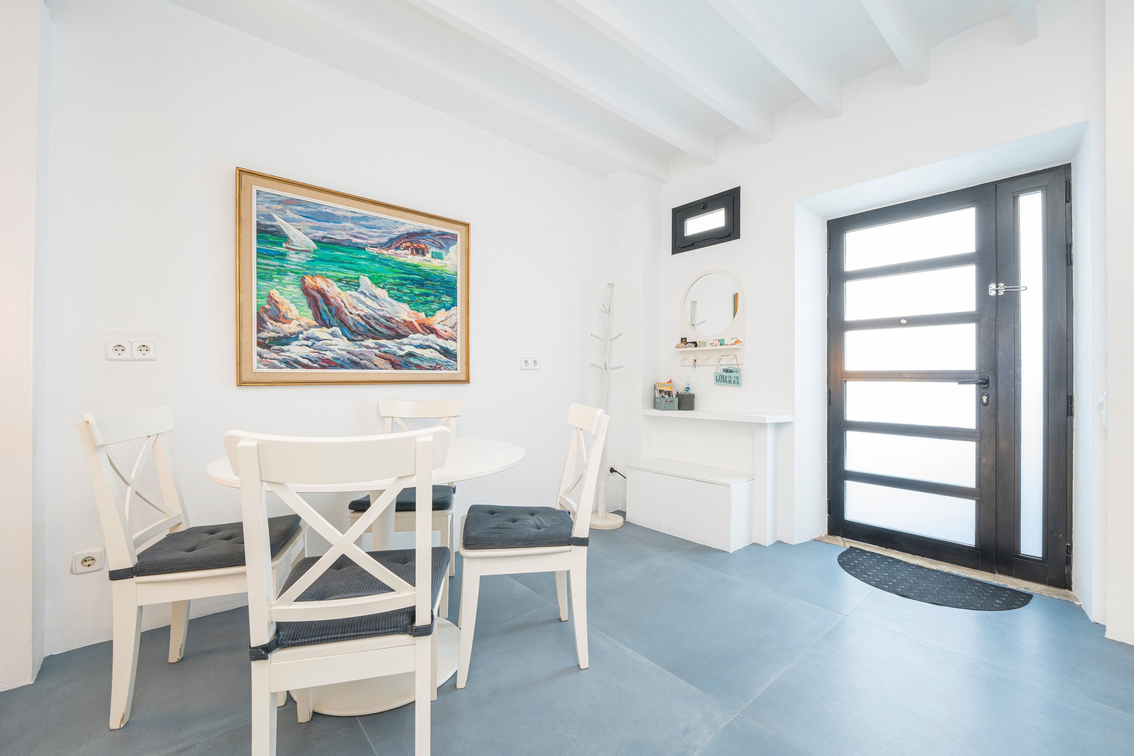 Un coin repas avec une table ronde et des chaises blanches, un tableau au mur, une porte avec des panneaux en verre dépoli et une petite étagère avec quelques objets.