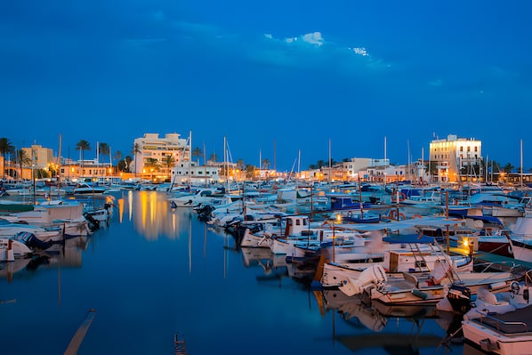 A marina at dusk with numerous boats docked, waterfront buildings, and palm trees.