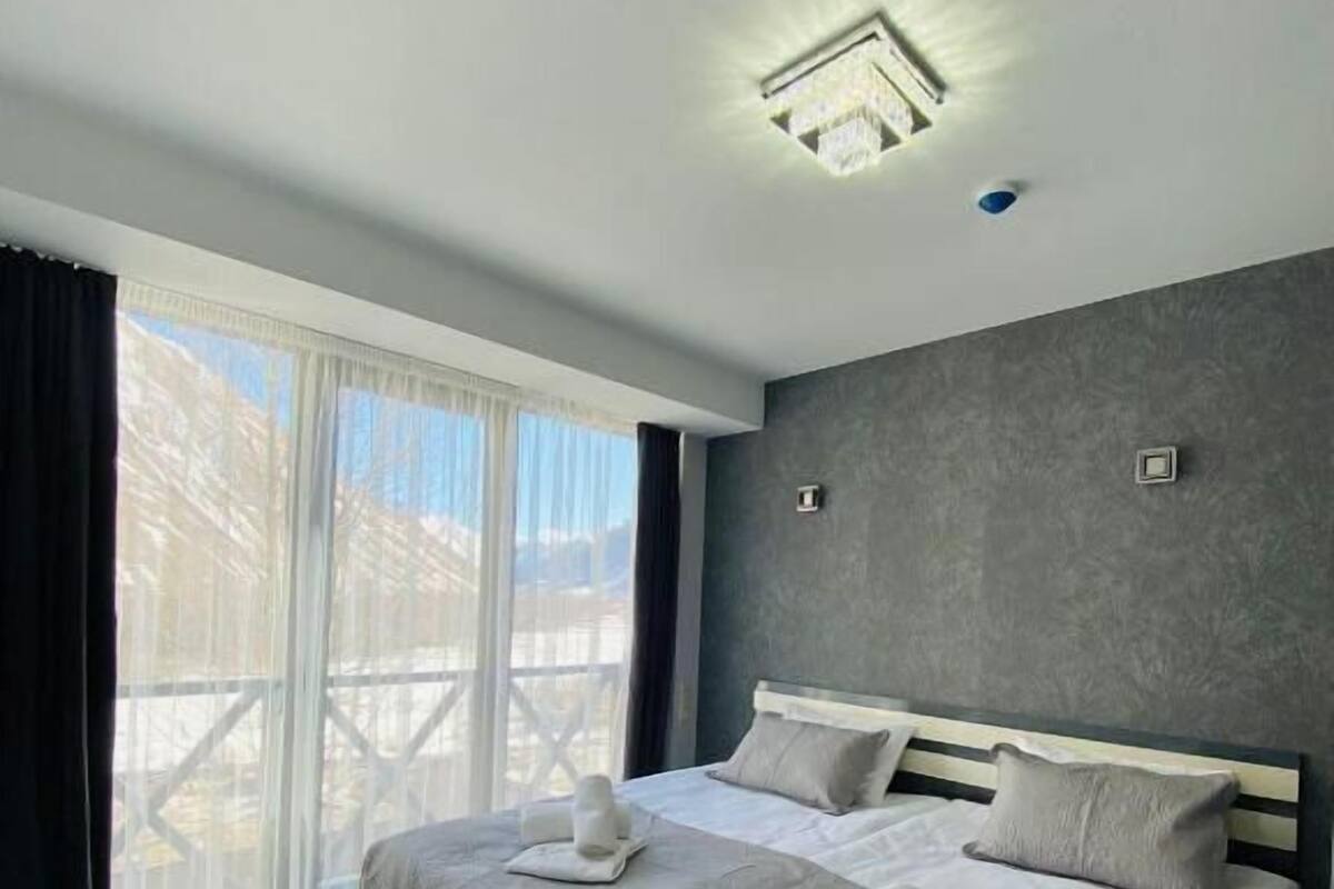 A modern bedroom with a large bed, a view of the outdoors through a sliding door, and a ceiling light fixture.