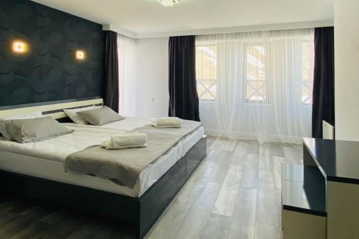 A modern hotel room with a large bed, a desk, and a view of the outdoors through sheer curtains.