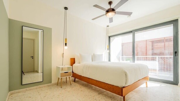 Een slaapkamer met een bed, een plafondventilator, een nachtkastje, een spiegel en een groot raam.