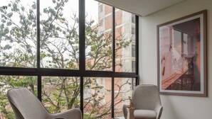 Ein Zimmer mit zwei Stühlen vor einem großen Fenster, einem runden Tisch mit Buch und Vase sowie einem gerahmten Bild an der Wand.