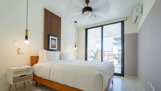 Een moderne slaapkamer met een houten bed, witte beddengoed, een nachtkastje, een plafondventilator en een groot raam met uitzicht op gebouwen.