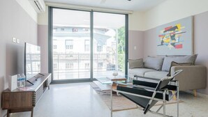 Een moderne woonkamer met een bank, een glazen salontafel, een televisie en een groot raam met uitzicht op gebouwen.