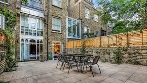 Une cour avec une table et des chaises, une clôture en bois et un bâtiment en briques doté de grandes fenêtres.