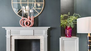 Une pièce avec une cheminée blanche, un miroir doté d’un cadre géométrique, une sculpture en corde rose, une plante en pot et une lampe.