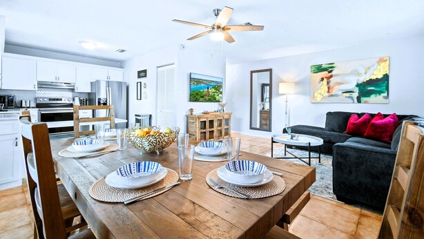 Una cocina moderna con armarios blancos, una mesa de comedor con platos azules y blancos, y un sofá negro en la sala.