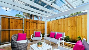 Un patio al aire libre con sillas de mimbre y mesas blancas, cercas de madera y techo de cristal.