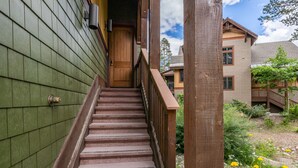 Una escalera de madera conduce a una puerta de madera, rodeada de follaje y un cielo despejado.