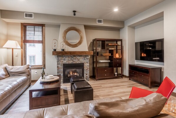 A living room with a fireplace, leather sofa, wooden coffee table, and a TV mounted on the wall. - Woodwinds 32 | 2 Private Decks | Hot Tub (Mammoth Lakes)