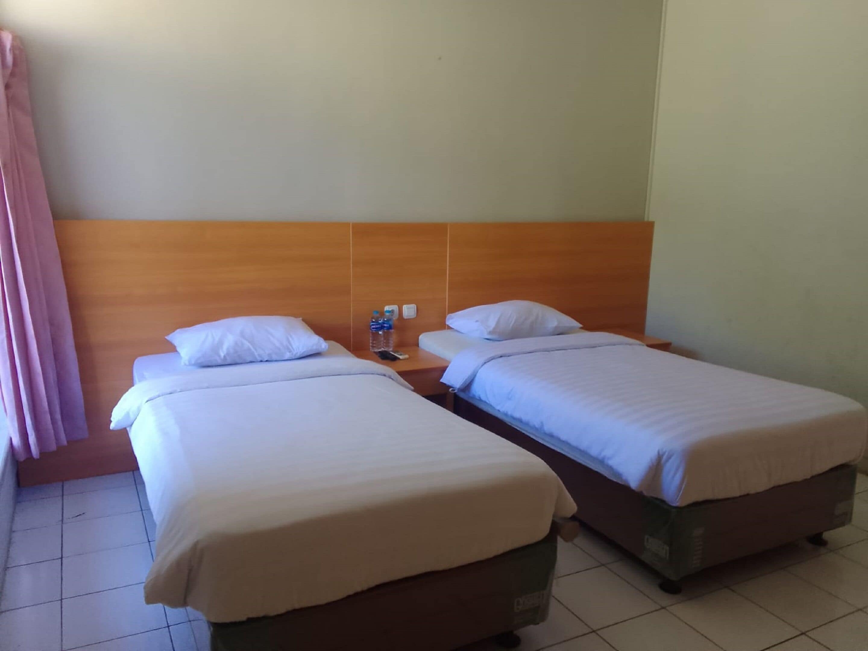 A hotel room with two single beds, a wooden headboard, a nightstand with a water bottle, and a curtain on the left.