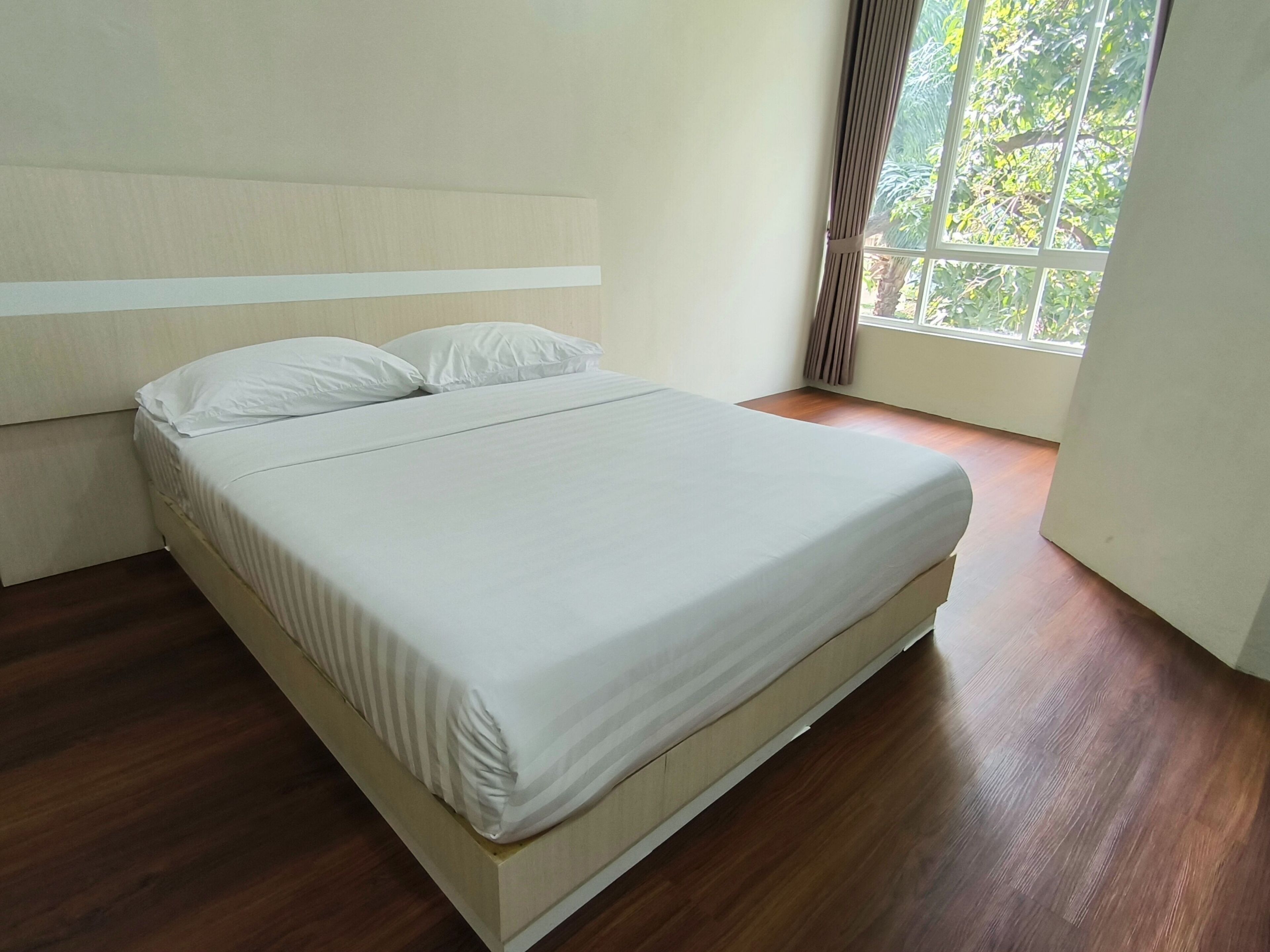 A neatly made bed in a room with a wooden floor and a window with curtains.