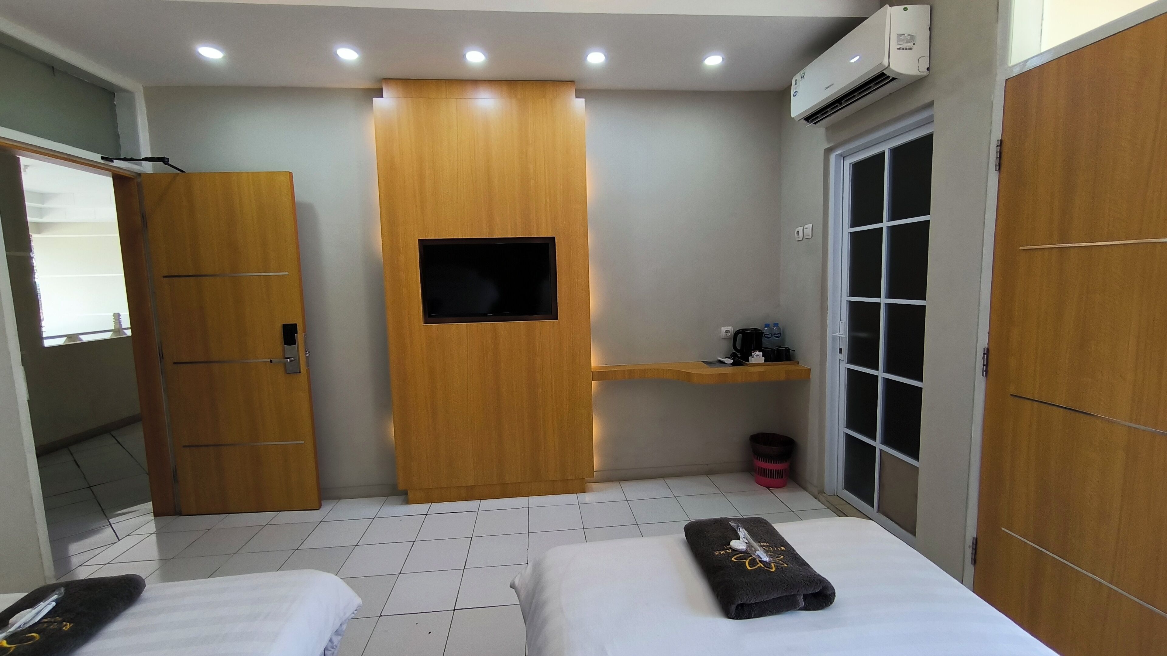 A hotel room with two beds, a wooden wardrobe, a small TV, a shelf with toiletries, and an air conditioning unit.
