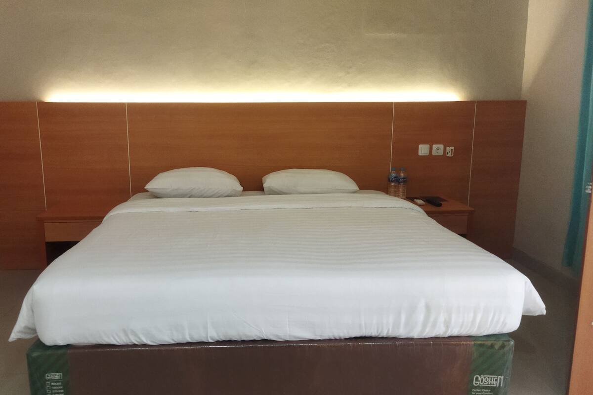 A hotel room with a large bed, wooden headboard, white bedding, two pillows, a nightstand with a remote control, and a water bottle.