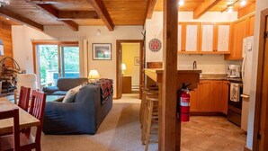 A cozy living room with a sofa, a dining table, and chairs. There is a kitchen area with wooden cabinets and a fire extinguisher. - Charming Okemo Home: Hot Tub, Fireplace, Sleeps 9 (Ludlow)