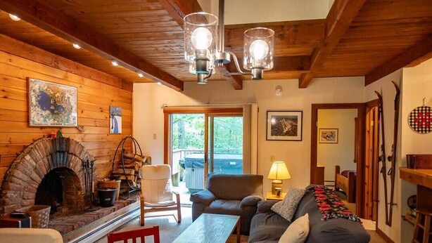 A cozy living room with a brick fireplace, wooden beams, a sofa, a table, and a lamp. - Charming Okemo Home: Hot Tub, Fireplace, Sleeps 9 (Ludlow)