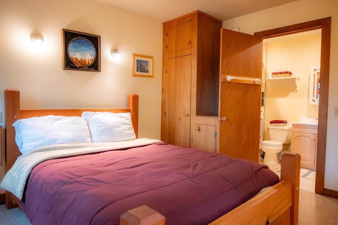 Habitació amb llit de fusta, un armari i un bany visible a través d'una porta oberta.
