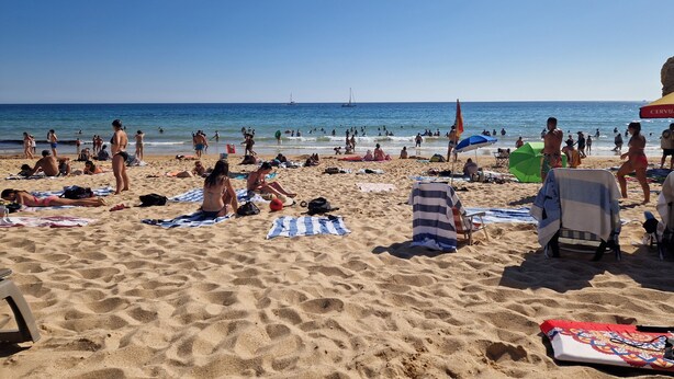 Ein Strand mit Menschen, die sich sonnen und schwimmen, ein Segelboot in der Ferne und ein Strandsonnenschirm.