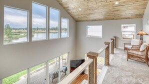 Ein geräumiges Zimmer mit großen Fenstern, das Blick auf einen See und Grünflächen bietet, mit Holzbalkendecke und einer gemütlichen Sitzecke mit Korbmöbeln.