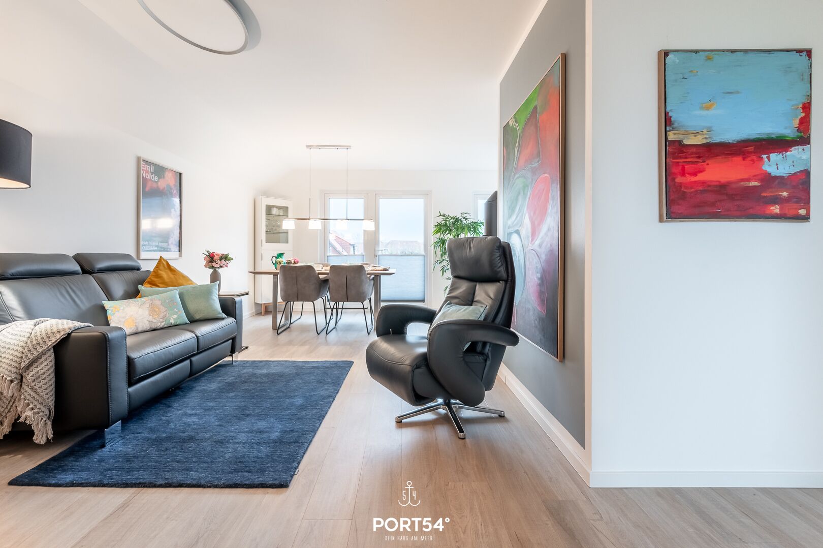 A modern living room with a black leather sofa, a black office chair, a dining table with chairs, and a large abstract painting on the wall.