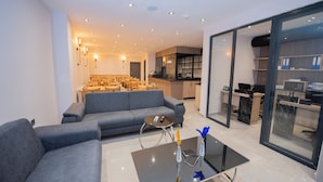 A modern living room with a grey sofa, a glass coffee table, and a kitchen area visible through glass doors. - Mahya Hotel (Istanbul)
