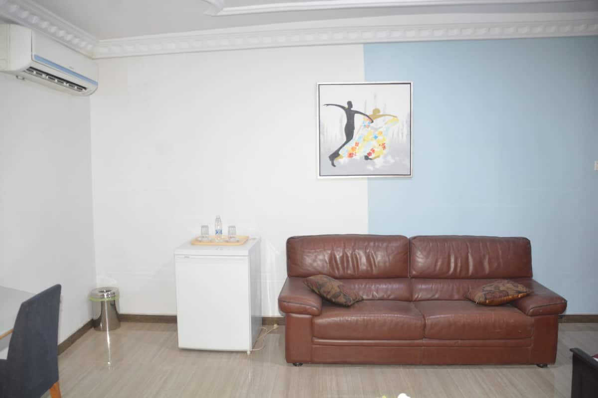 Una sala de estar con un sofá de cuero marrón, un mini refrigerador blanco y una obra de arte enmarcada en la pared.