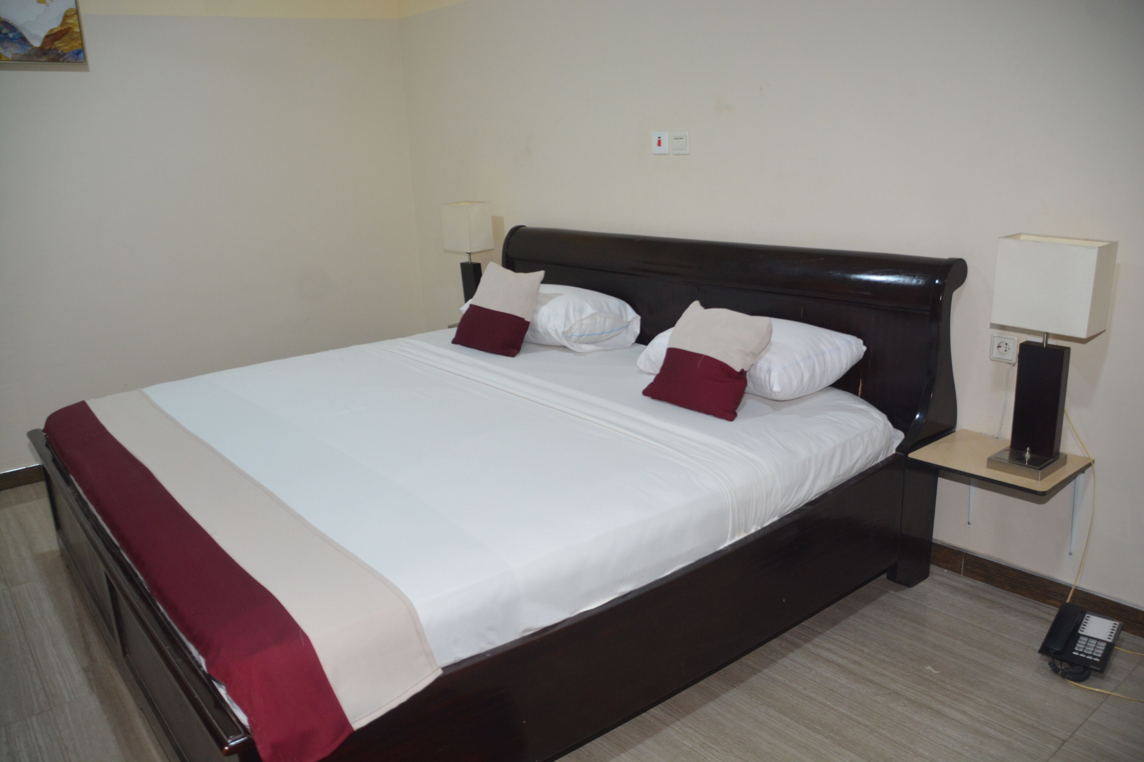 Cama bem arrumada com roupa de cama branca e bordô, cabeceira escura e mesa de cabeceira com abajur e telefone.