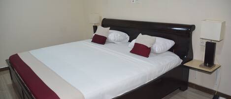 Cama bem arrumada com roupa de cama branca e bordô, cabeceira escura e mesa de cabeceira com abajur e telefone.
