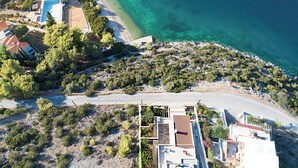 Ett flygbildsbild av ett kustområde med en strand, klart blått vatten och bostadshus med synliga tak och andra byggnadsstrukturer.