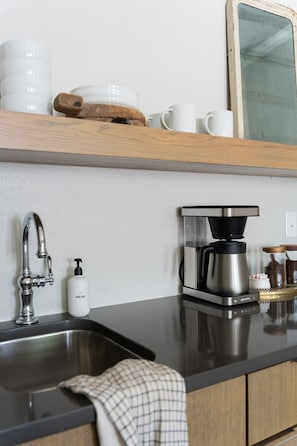 Una cocina con un lavamanos de acero inoxidable, una cafetera y un estante de madera con tazas y un tarro.