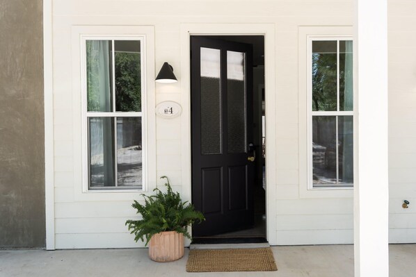 A white house with a black front door, two windows, a potted plant, and a wall-mounted light fixture.