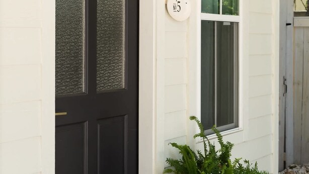 A black door with frosted glass panels, a white house number plaque, a potted plant, and a wall-mounted light fixture. - Cozy Modern Suite with Hot Tub (Fredericksburg)