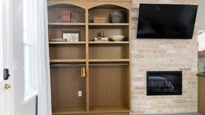 A room with a wooden shelving unit, a flat-screen TV, and a fireplace. - Cozy Modern Suite with Hot Tub (Fredericksburg)