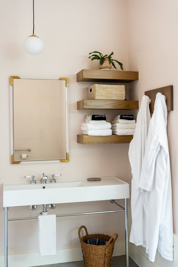 A bathroom with a white sink, a mirror with a gold frame, wooden shelves, a hanging robe, and a wicker basket.