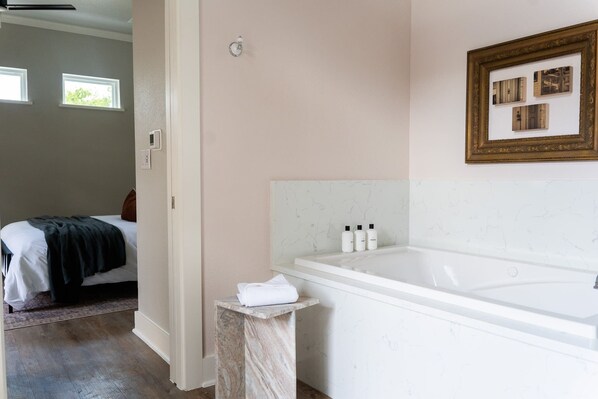 Une salle de bain avec une grande baignoire, un lit recouvert d’une couverture sombre et une petite table avec des articles de toilette.