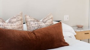 Un lit bien fait, agrémenté d’oreillers à motifs et d’un coussin marron, un luminaire fixé au mur et une petite table de chevet avec un bol.