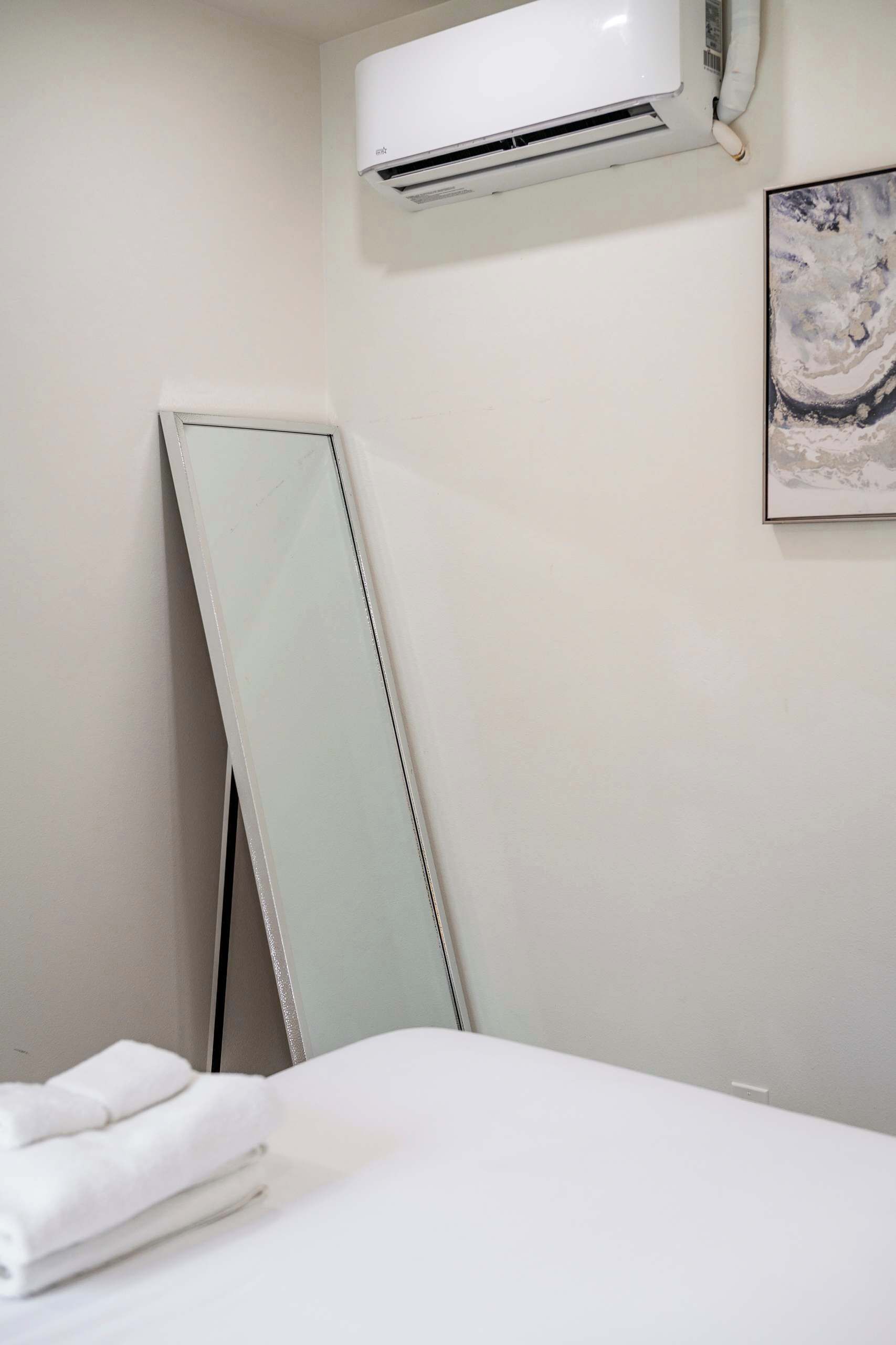 Une chambre d’hôtel avec un lit, un grand miroir, un appareil de climatisation et une œuvre d’art encadrée accrochée au mur.