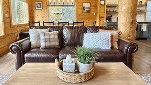 A cozy cabin interior with a leather sofa, a wooden dining table, and a large window. - Cozy Cabin Close to Lake Cascade w/ Hot Tub (Cascade)