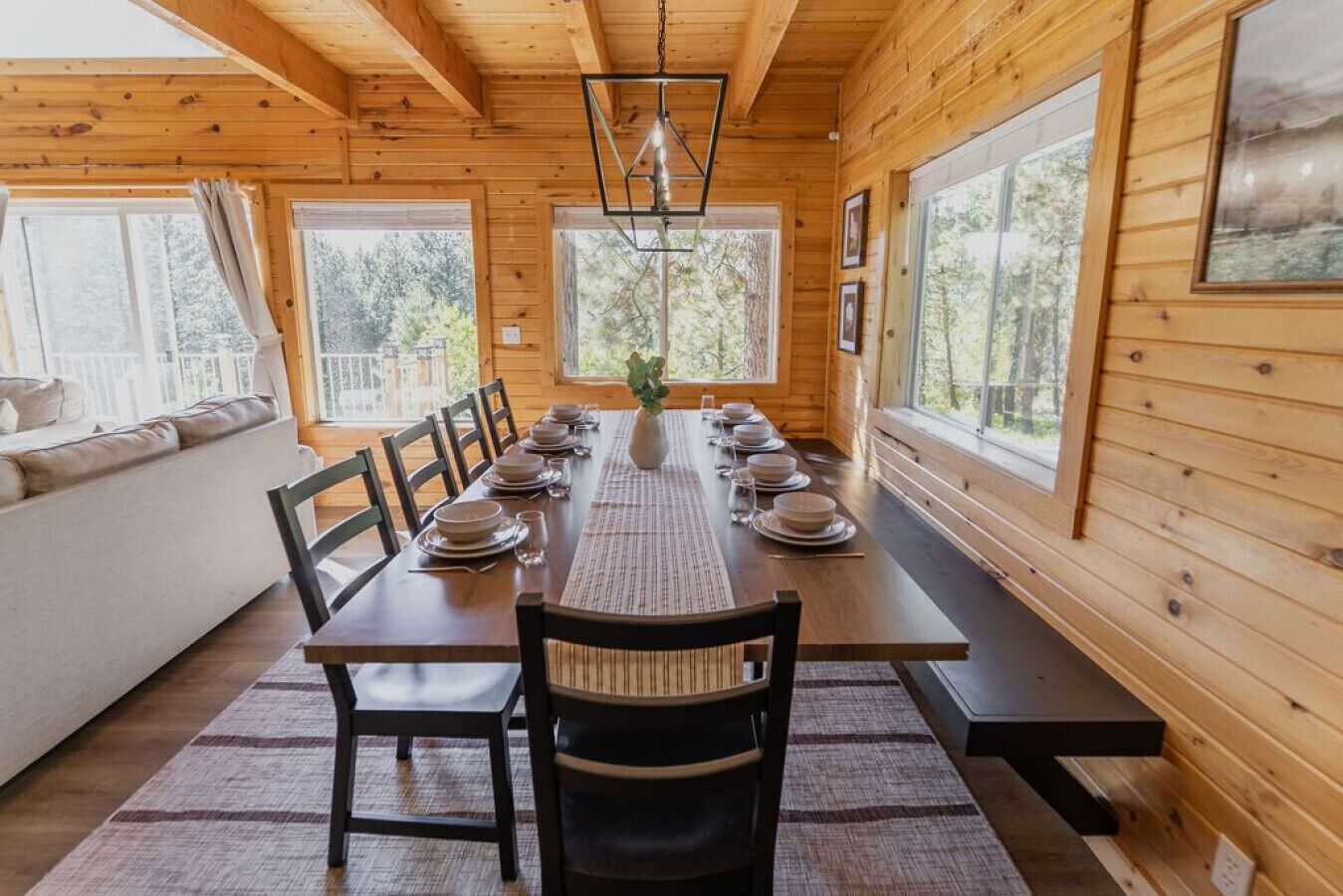 Une salle à manger aménagée dans une cabane en bois, avec une longue table dressée pour six personnes, de grandes fenêtres et une vue sur la forêt.