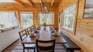 A wooden cabin dining area with a long table set for six, large windows, and a view of the forest. - Cozy Cabin Close to Lake Cascade w/ Hot Tub (Cascade)