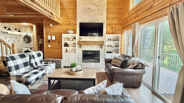 A cozy living room with a stone fireplace, wooden walls, and large windows. - Cozy Cabin Close to Lake Cascade w/ Hot Tub (Cascade)