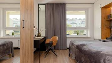 A modern bedroom with two beds, a desk, and a chair. There are large windows with curtains, a view of buildings outside, and a wooden door.