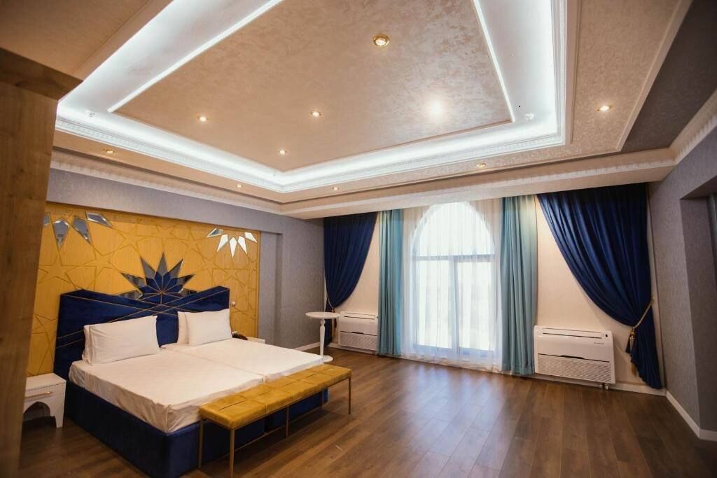 A spacious bedroom with a large bed, a wooden bench, a ceiling light fixture, and a window with curtains.
