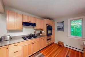 A compact kitchen with wooden cabinets, a gas stove, a toaster, and a window with a view of greenery.