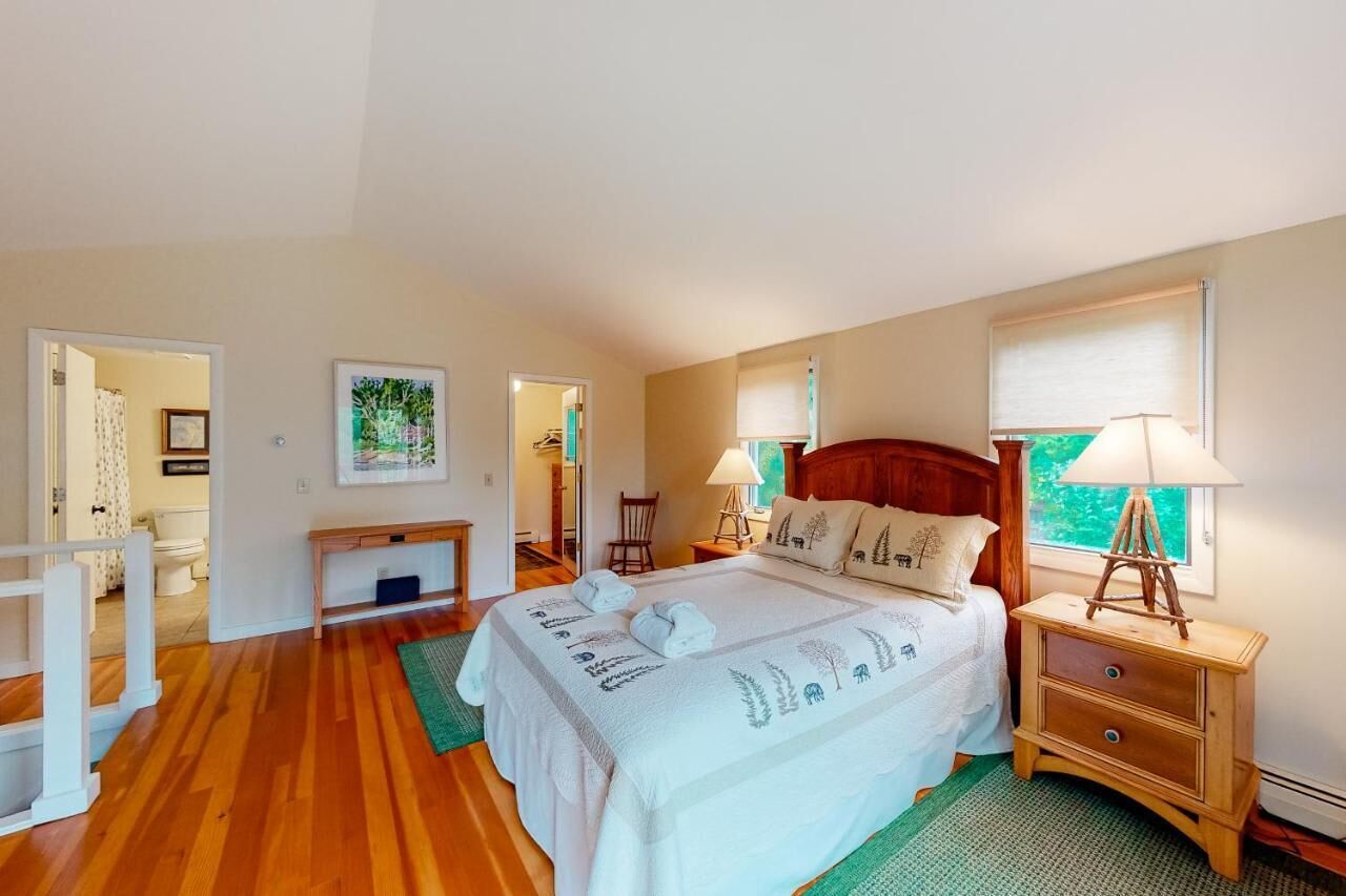 A bedroom with a bed, bedside tables, a nightstand, a window with blinds, and a door leading to another room.