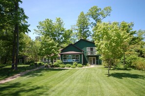 Une maison verte avec une grande fenêtre et un balcon, entourée d’une pelouse bien entretenue et d’arbres.