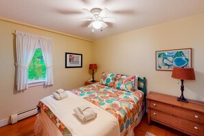 Une chambre à coucher comprenant un lit, un ventilateur de plafond, une fenêtre avec des rideaux, une table de chevet, une lampe, une commode et deux tableaux encadrés.