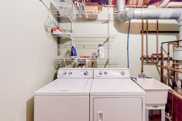 A laundry area with two front-loading washing machines, a metal shelf unit with storage, and visible plumbing.