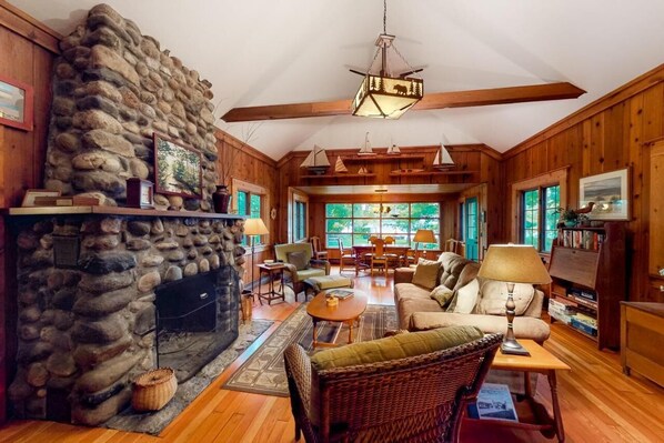 A cozy cabin interior with a stone fireplace, wooden walls, and a dining area with a view.