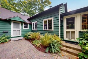 Une maison verte aux boiseries blanches, un chemin pavé de briques et diverses plantes.